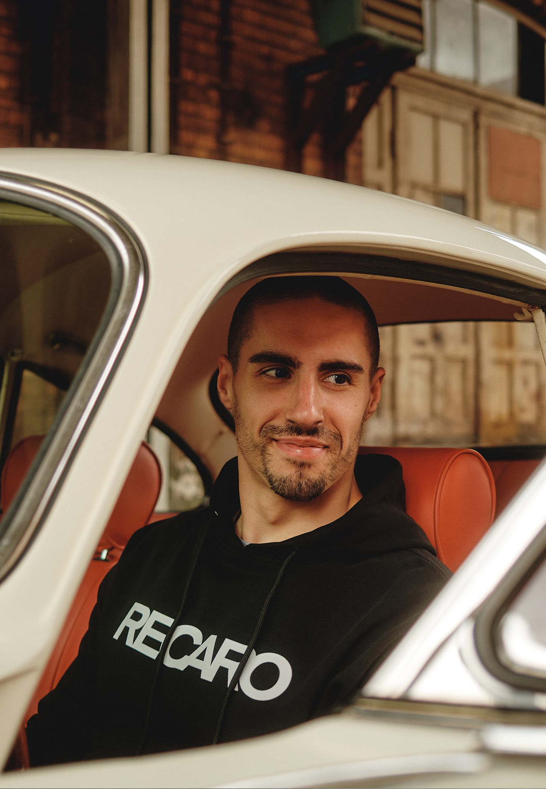 Ein Mann mit kurzen Haaren und gestutztem Bart sitzt in einem Auto mit orangefarbenen Sitzen, trägt den RECARO Shop Hoodie Originals aus entspannt geschnittenem Baumwollmix, schaut aus dem offenen Fenster und lächelt leicht.