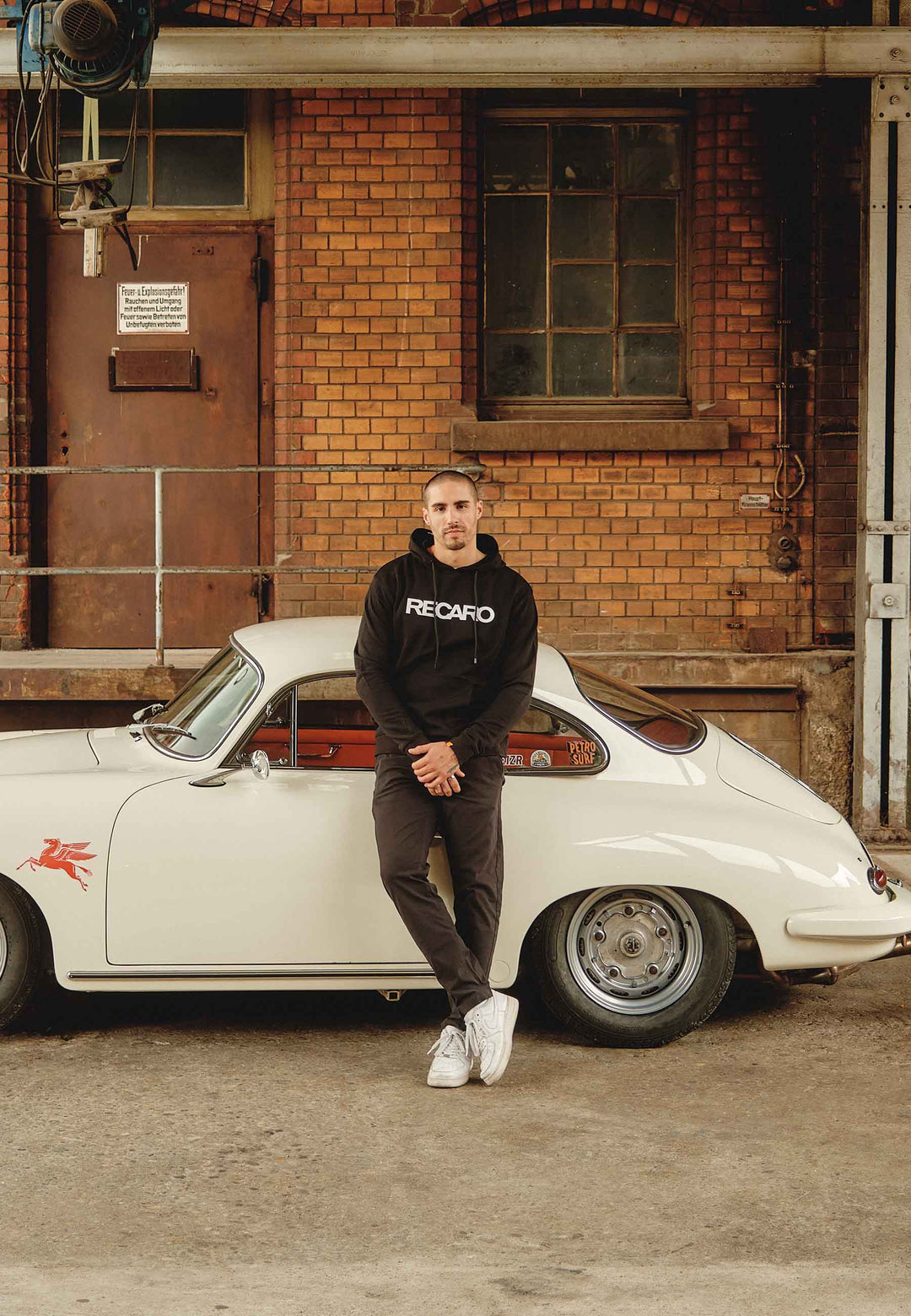 Ein Mann in einem Relaxed-Fit Hoodie Originals aus dem RECARO Shop und weißen Turnschuhen lehnt an einem weißen Oldtimer, der neben einem Backsteingebäude mit Industrieelementen geparkt ist.