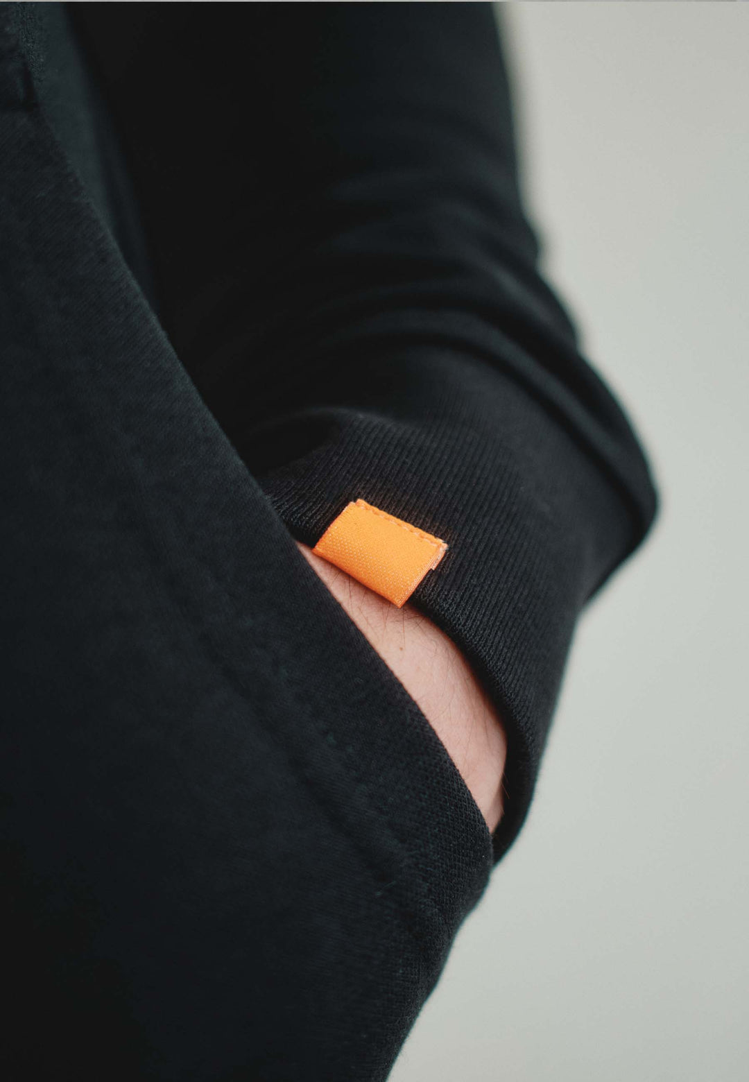 Eine Nahaufnahme einer Hand in der Tasche eines schwarzen Hoodie Originals aus dem RECARO Shop, mit einem kleinen orangefarbenen Stoffetikett, das in der Nähe der Taschennaht sichtbar ist.