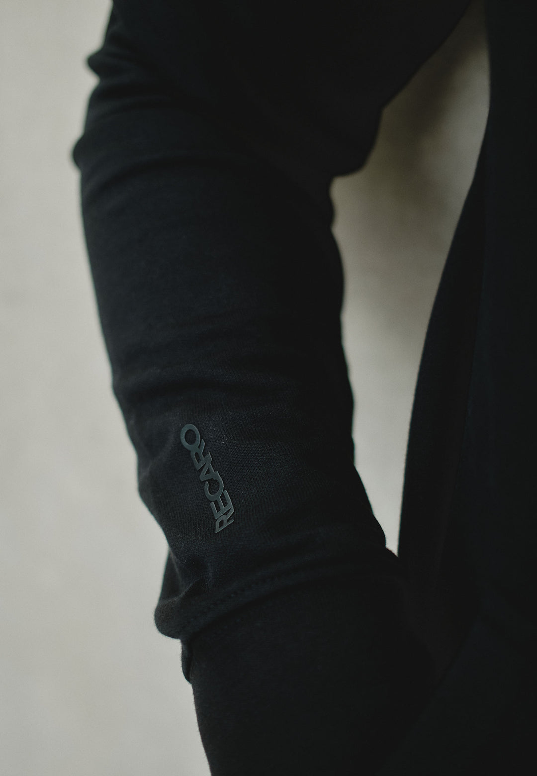 Eine Nahaufnahme eines Arms in einem locker sitzenden schwarzen Hoodie Originals aus dem RECARO Shop, aus weichem Baumwollmischgewebe, mit dem Aufdruck „RECARO“ in kleinen, hellen Buchstaben auf dem Ärmel.
