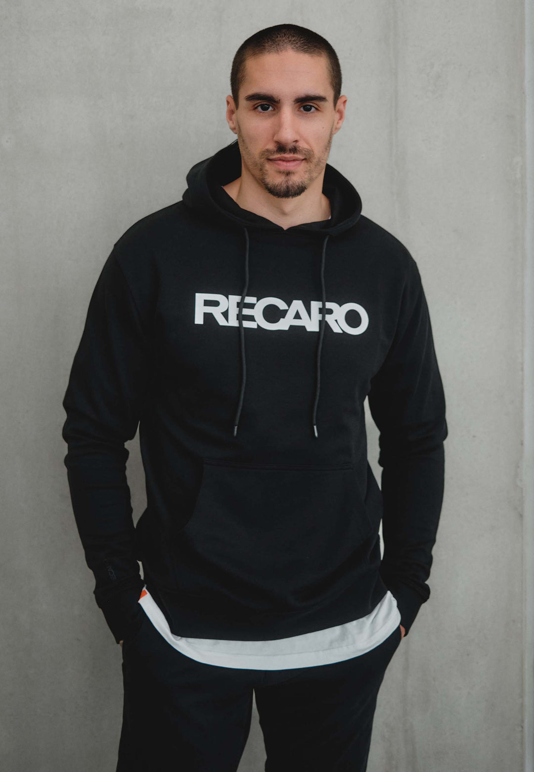 Ein Mann mit kurzen Haaren und gestutztem Bart steht vor einer schlichten hellgrauen Wand. Er trägt den Hoodie Originals aus dem RECARO Shop. Er ist aus weichem Baumwollmischgewebe gefertigt, hat die Hände in den Hosentaschen und blickt in die Kamera.