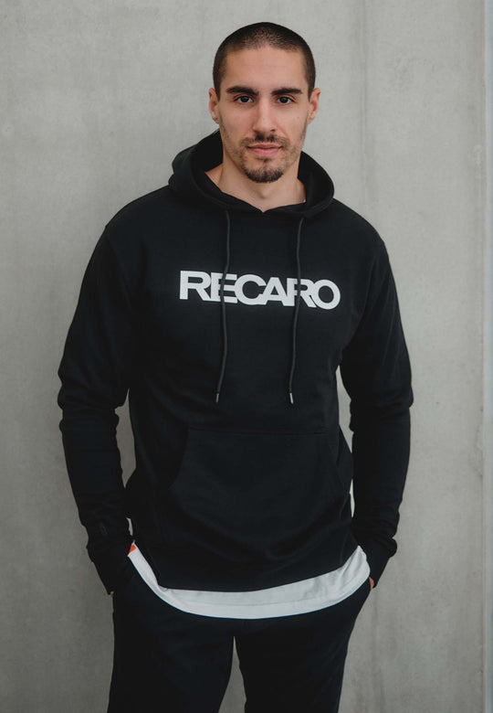 Ein Mann mit kurzen Haaren und gestutztem Bart steht vor einer schlichten hellgrauen Wand. Er trägt den Hoodie Originals aus dem RECARO Shop. Er ist aus weichem Baumwollmischgewebe gefertigt, hat die Hände in den Hosentaschen und blickt in die Kamera.