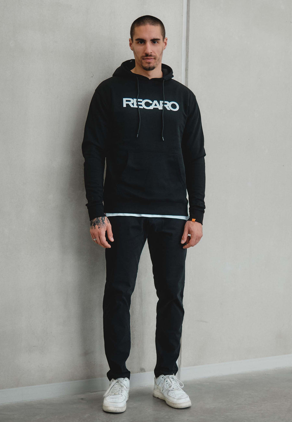 Ein Mann mit kurzen Haaren und Handtattoos steht vor einer schlichten hellen Wand und trägt einen Hoodie Originals aus dem RECARO Shop, kombiniert mit einer schwarzen Hose und weißen Turnschuhen.