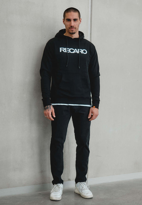 Ein Mann mit kurzen Haaren und Handtattoos steht vor einer schlichten hellen Wand und trägt einen Hoodie Originals aus dem RECARO Shop, kombiniert mit einer schwarzen Hose und weißen Turnschuhen.