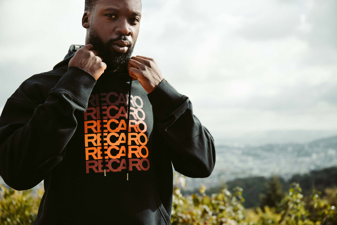 Ein Mann steht im Freien und hält den Kragen seines schwarzen Hoodie Spektrum von RECARO, auf dem RECARO in wiederholten Farbverläufen in Rot und Orange für einen Retro-Look abgebildet ist. Verschwommenes Grün und ein bewölkter Himmel bilden den Hintergrund.