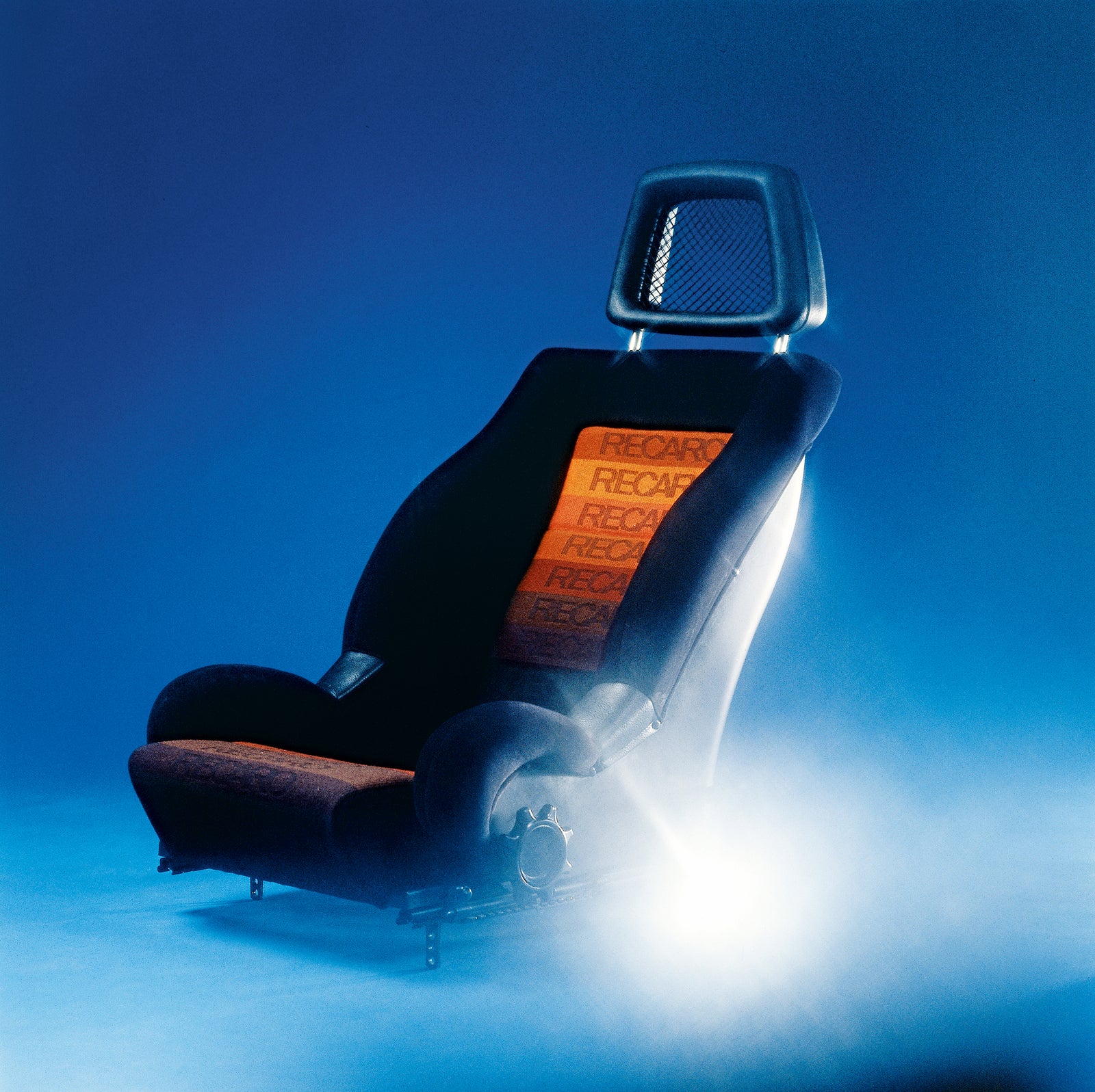 Ein sportlicher Recaro-Autositz mit schwarz-oranger Polsterung, auf dem das Recaro-Logo wiederholt aufgedruckt ist, wird vor einem blauen, nebligen Hintergrund beleuchtet.