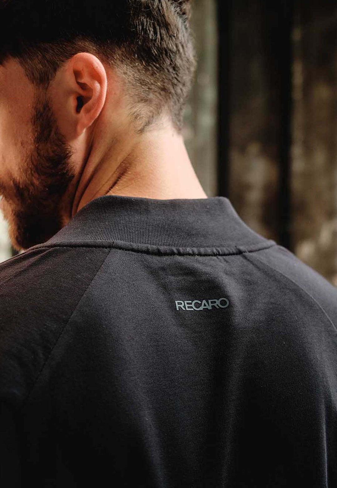 Eine Nahaufnahme zeigt Rücken und Nacken eines Mannes, der einen dunklen RECARO_Merchandising Sweat Blouson mit "RECARO" in kleinen weißen Buchstaben unterhalb des Kragens trägt. Der weiche Baumwoll-Sweat-Stoff hebt sich von dem unscharfen Hintergrund ab.