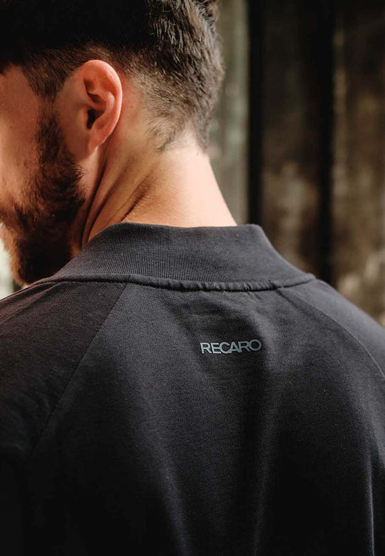 Eine Nahaufnahme zeigt Rücken und Nacken eines Mannes, der einen dunklen RECARO_Merchandising Sweat Blouson mit "RECARO" in kleinen weißen Buchstaben unterhalb des Kragens trägt. Der weiche Baumwoll-Sweat-Stoff hebt sich von dem unscharfen Hintergrund ab.