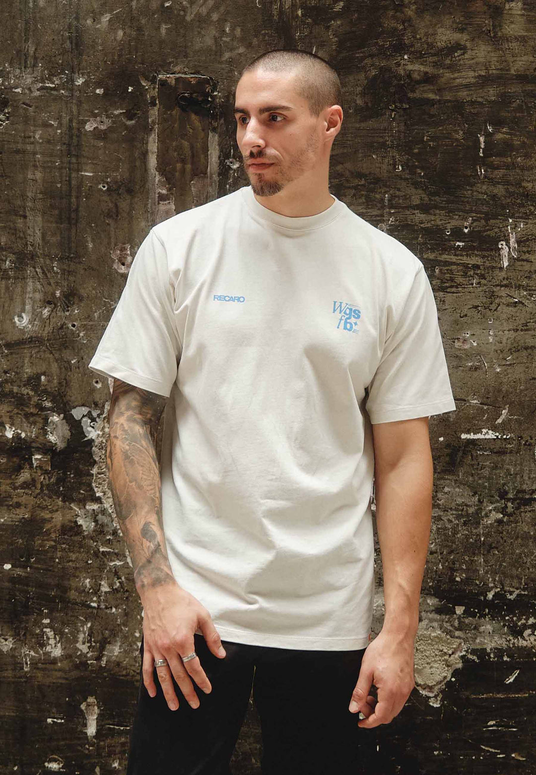 Ein Mann mit kurzen Haaren und Tätowierungen auf dem linken Arm steht an einer strukturierten dunklen Wand und trägt das RECARO Oversized Shirt WGSFB – aus schwerer Baumwolle mit dezentem blauem Text – und eine dunkle Hose.