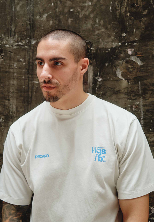 Ein Mann mit rasiertem Kopf, gestutztem Bart und Schnurrbart trägt ein cremefarbenes RECARO Oversized Shirt WGSFB mit blauem Text, steht vor einer strukturierten dunklen Betonwand und blickt leicht zur Seite.