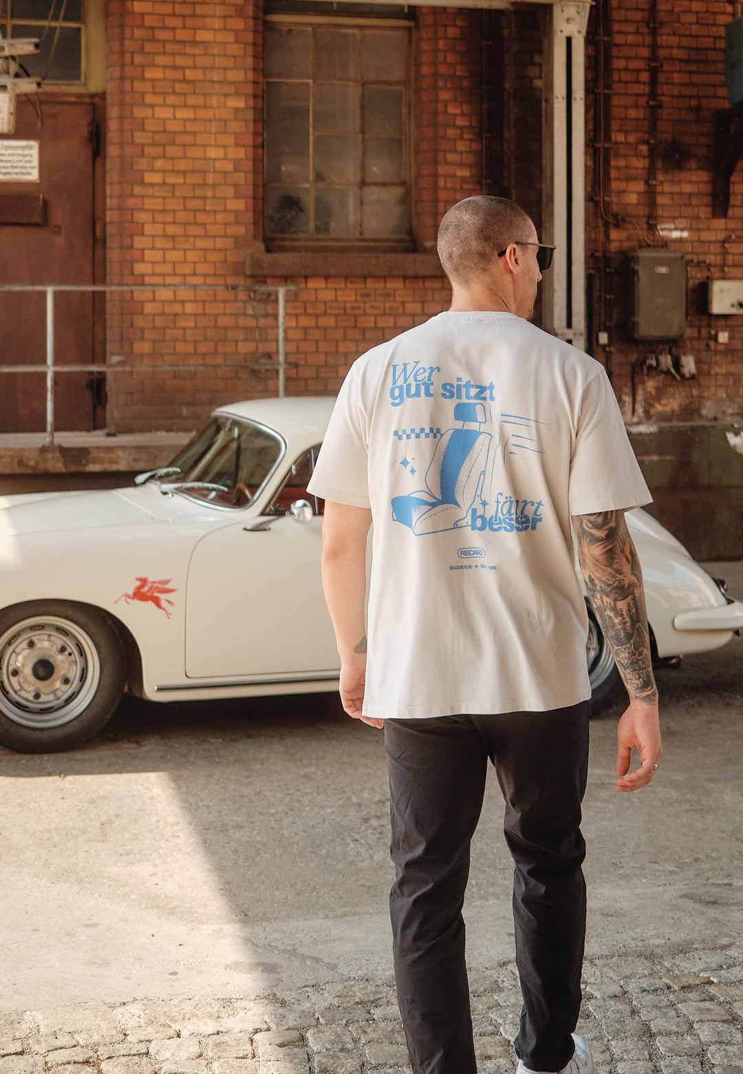 Ein Mann mit tätowiertem rechten Arm, Sonnenbrille und einem übergroßen beigen RECARO Oversized Shirt WGSFB mit blauer Schrift auf dem Rücken geht auf ein weißes Oldtimerauto zu, das vor einem Backsteingebäude geparkt ist.