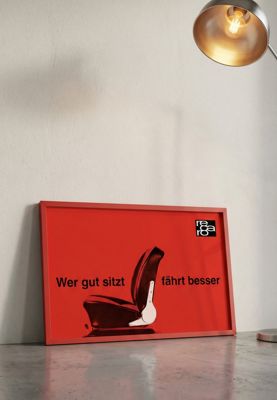 Das Poster "Wer gut sitzt fährt besser" aus dem RECARO Shop zeigt eine Autositz-Illustration, einen deutschen Text und ein weißes Logo auf schwarzem Untergrund oben rechts. Es hat einen roten Rahmen und ist an eine Wand gelehnt unter einer beleuchteten Lampe angebracht.