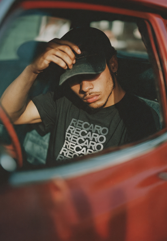 Eine Person sitzt in einem roten Auto und neigt die Krempe ihrer schwarzen Mütze. Ihr Recaro_Merch_Daniel T-Shirt Spektrum grey sorgt für einen Hauch von Retro, und die weißen Buchstaben fangen das Sonnenlicht durch das teilweise geöffnete Fenster ein.