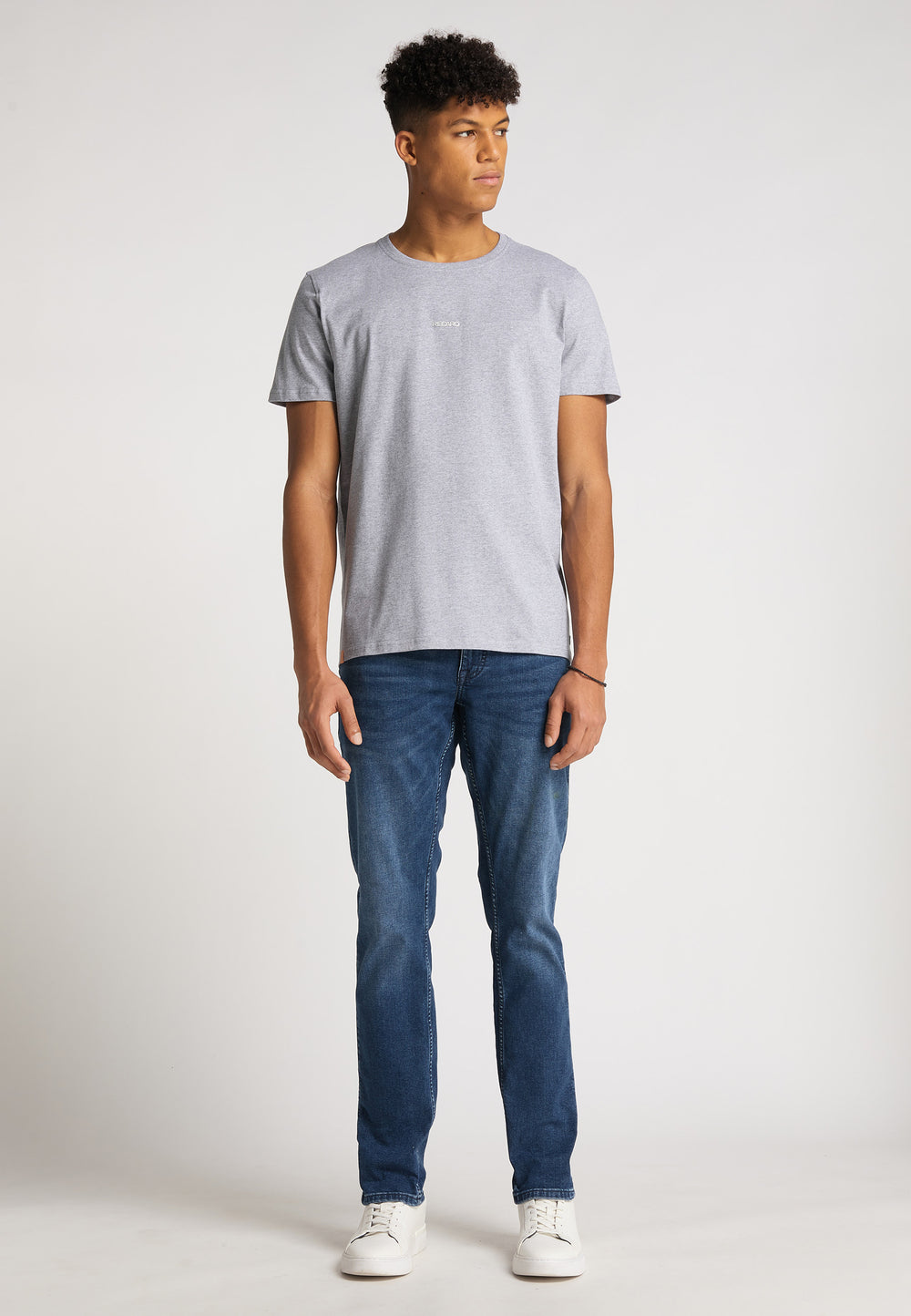 Ein junger Mann steht vor einem weißen Hintergrund. Er trägt das RECARO T-Shirt mit Rückenaufdruck in Hellgrau, dazu blaue Jeans und weiße Turnschuhe. Er blickt leicht nach links, die Arme hängen entspannt an den Seiten.