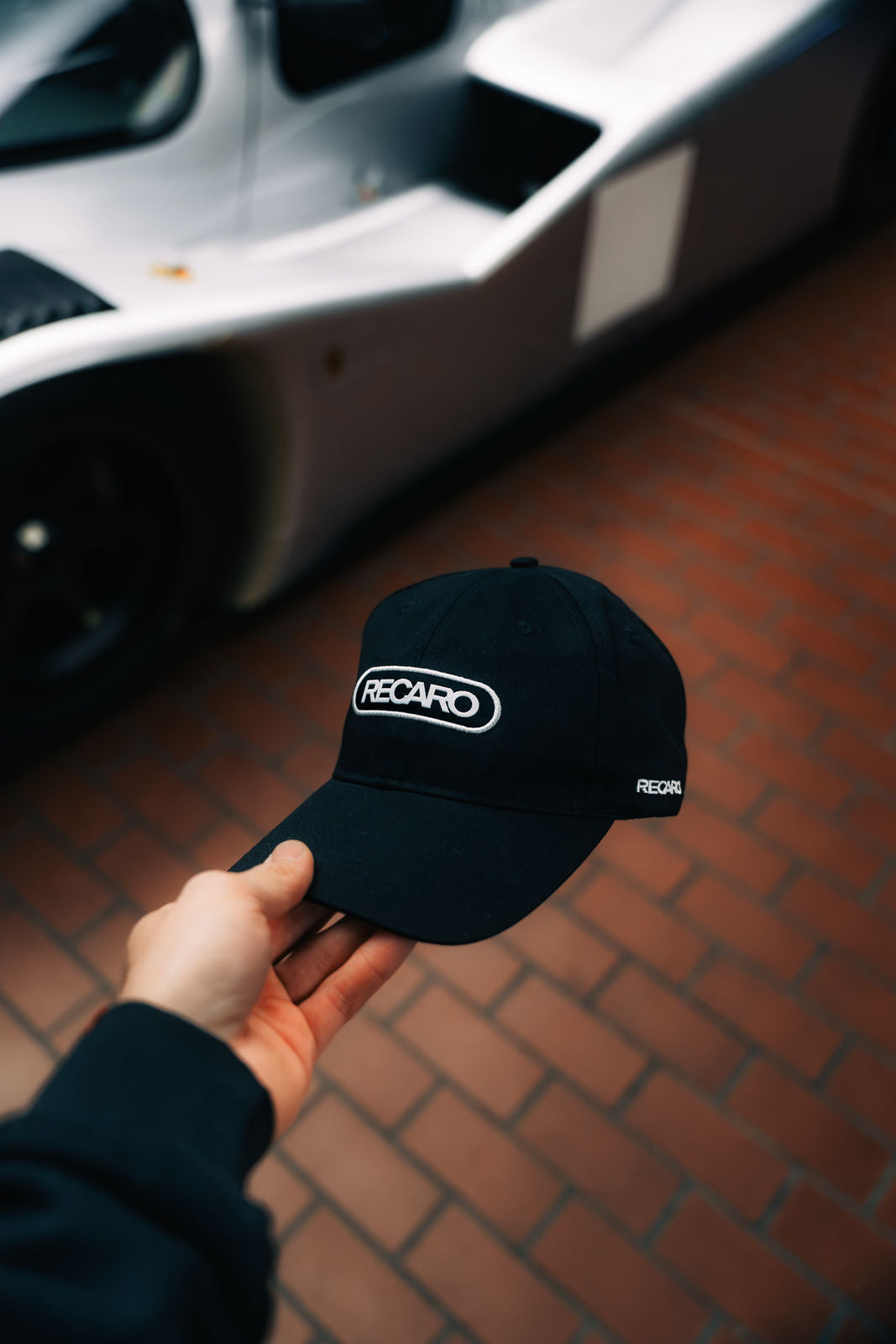 Eine Person hält eine RECARO Shop Baseball Cap Basic black vor einen silbernen Sportwagen, der auf rotbraunem Backsteinpflaster geparkt ist.