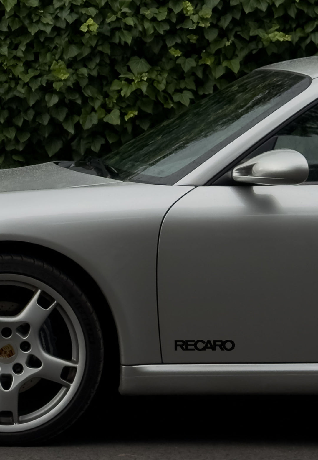 Nahaufnahme eines silbernen Sportwagens mit RECARO Shop Premium Vinyl Autoaufkleber (2 Stück) an der Seite, geparkt vor einer grünen Hecke. Die Aufnahme hebt den vorderen Kotflügel, das Rad, die Tür und den Spiegel hervor und zeigt die Wetterbeständigkeit des Vinyls.