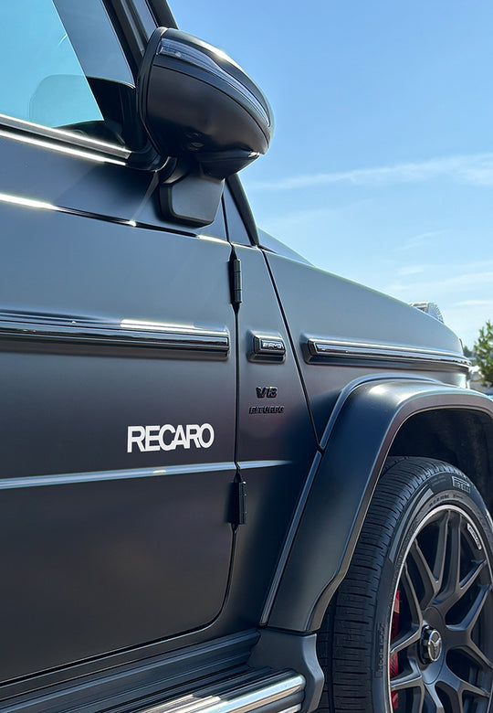Nahaufnahme eines schwarzen Mercedes-Benz G-Klasse SUV mit RECARO Shop Premium Vinyl Autoaufkleber (2 Stück) auf der Tür, V8 Biturbo-Plakette und auffälligen schwarzen Leichtmetallrädern unter strahlend blauem Himmel.