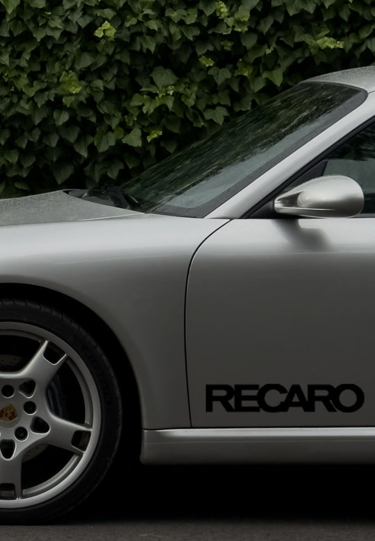 Seitenansicht eines silbernen Sportwagens mit RECARO Premium Vinyl Autoaufkleber (2 Stück) aus dem RECARO Shop an der Tür, Porsche Rad sichtbar, grüner Blätterhintergrund. Der Aufkleber bietet stilvolle Optik und Witterungsbeständigkeit.