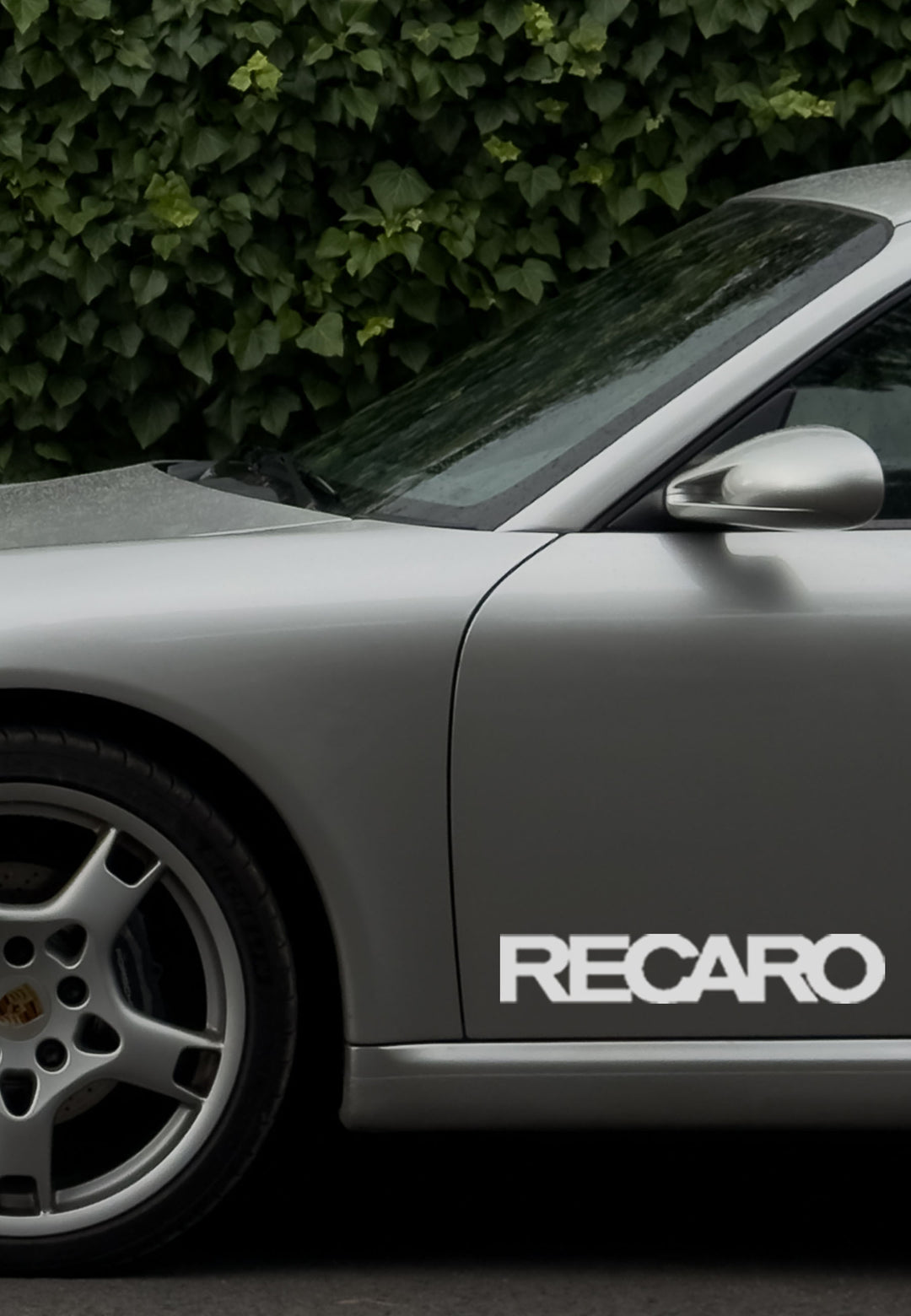 Seitenansicht eines silbernen Sportwagens mit einem RECARO Premium Vinyl Autoaufkleber (2 Stück) aus dem RECARO Shop auf der Tür, der die witterungsbeständige Oberfläche und das detaillierte Porsche-Rad hervorhebt, geparkt zwischen grünen Büschen.