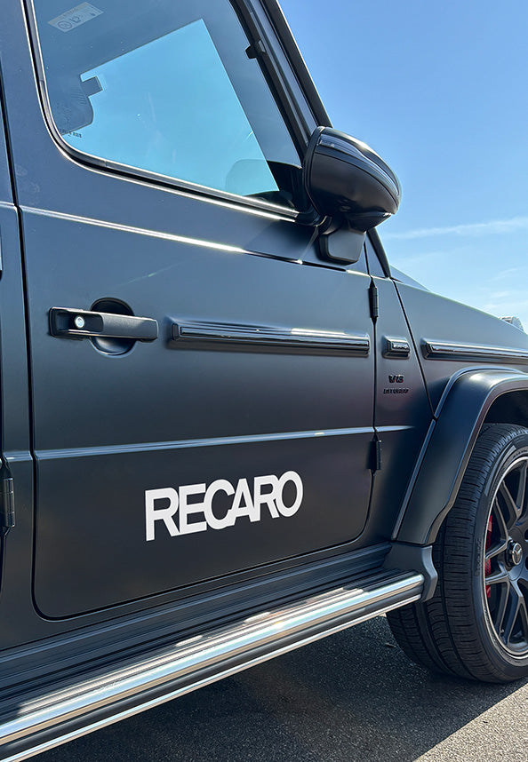 Ein mattschwarzer Mercedes-Benz G-Klasse SUV trägt den RECARO Premium Vinyl Autoaufkleber (2 Stück) von RECARO Shop auf der Tür, mit sichtbarer V8 Biturbo-Plakette und schwarzem Leichtmetallrad unter strahlend blauem Himmel.