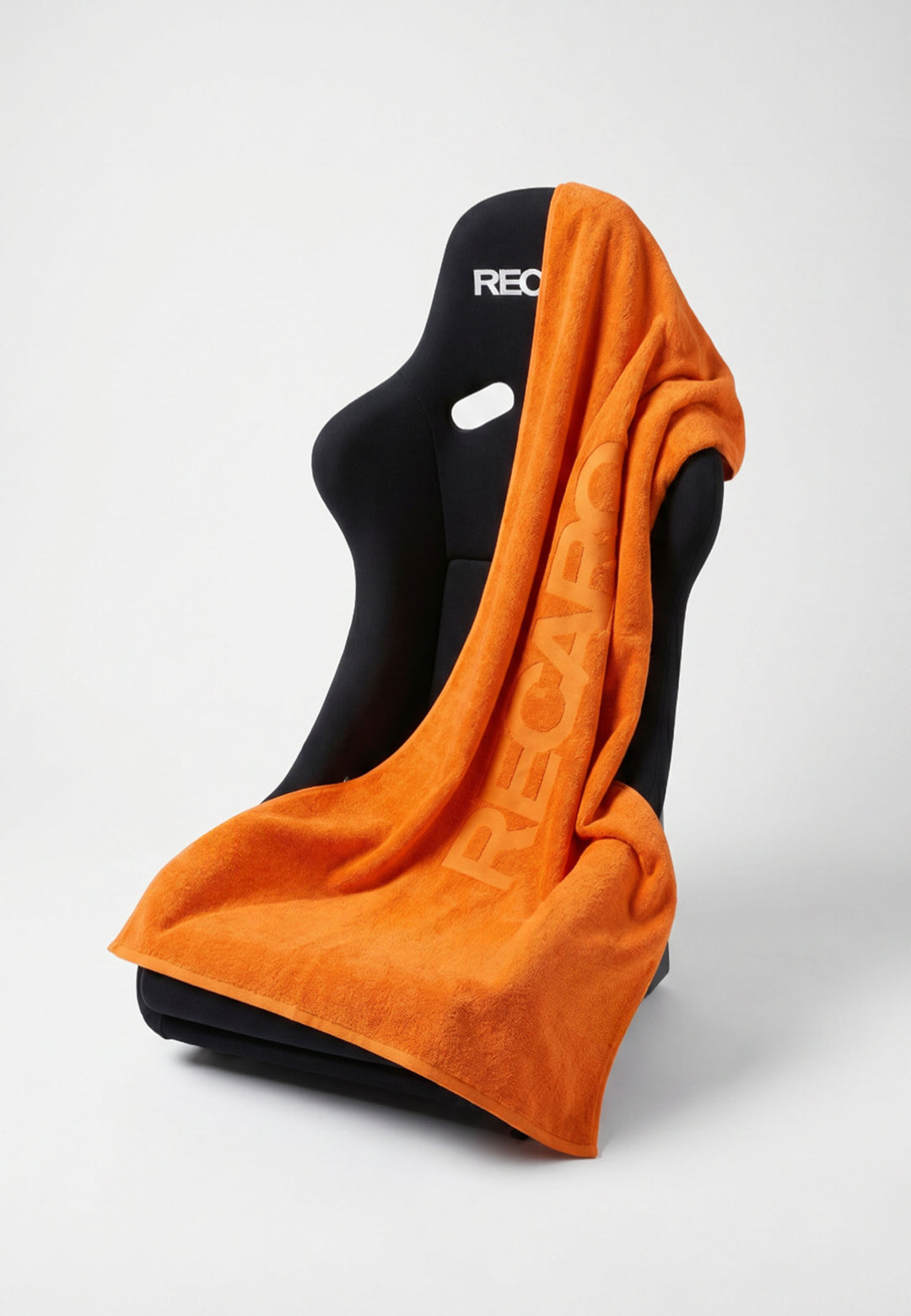 Ein schwarzer Rennsport-Sitz ist mit dem RECARO XXL Handtuch aus dem RECARO Shop in leuchtendem Orange drapiert. Das aus 100 % Baumwolle gefertigte Handtuch ist mit einem großen, geprägten RECARO-Logo versehen und wird vor einem schlichten weißen Hintergrund ansprechend über dem Stuhl präsentiert.