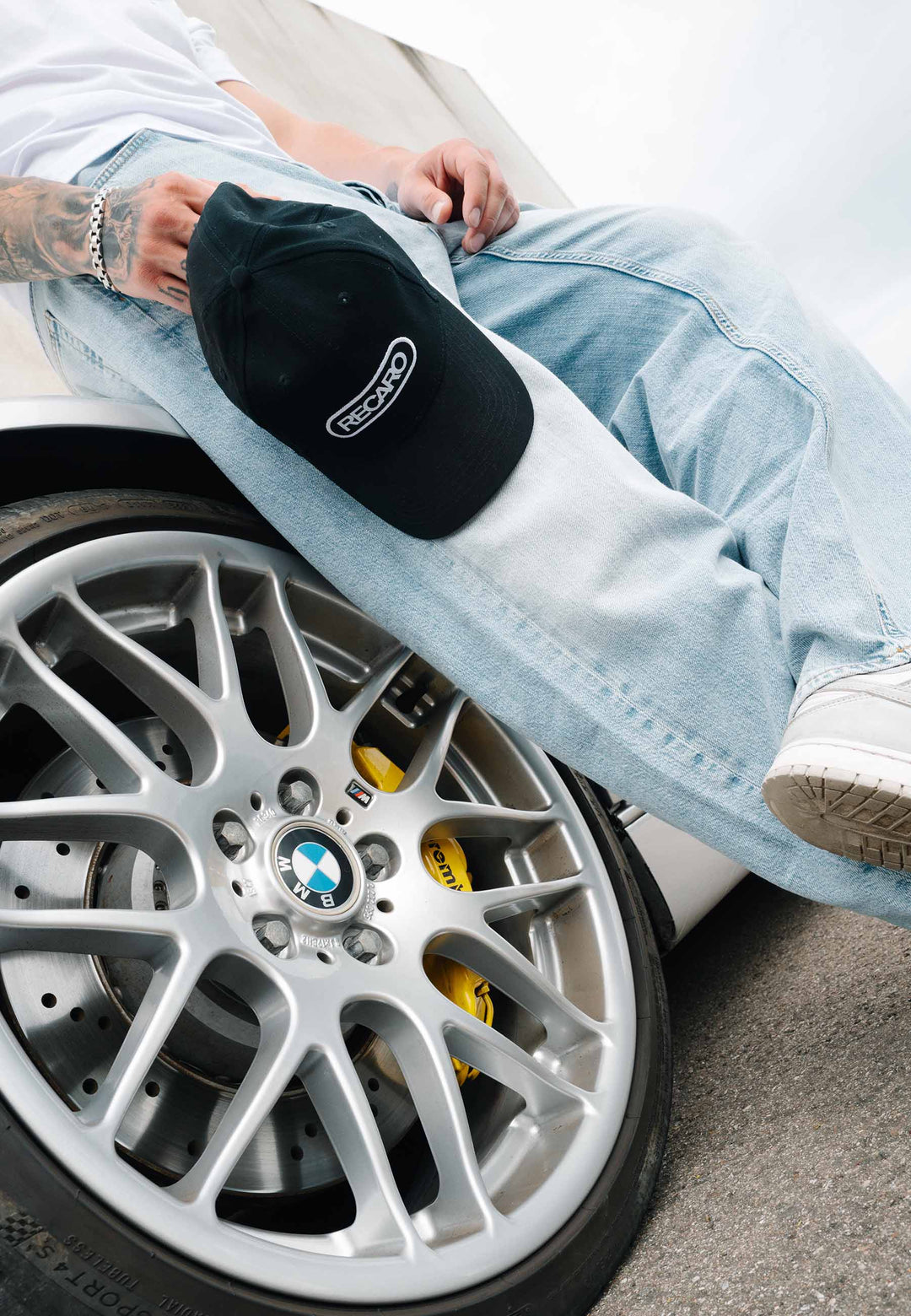 Eine Person in hellblauen Jeans und weißen Sneakers lehnt an einem weißen BMW und hält eine schwarze RECARO Shop Baseball Cap Basic in der Nähe des silbernen Leichtmetallrads mit den gelben Bremssätteln in der Hand.