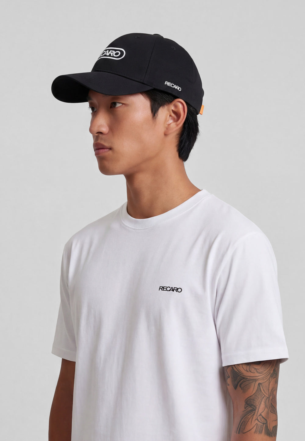 Mit einer schwarzen RECARO Shop Baseball Cap Basic und einem weißen Baumwolle T-Shirt, beides mit RECARO Logo, steht eine Person mit kurzen dunklen Haaren und einer Armtätowierung vor einem schlichten grauen Hintergrund.