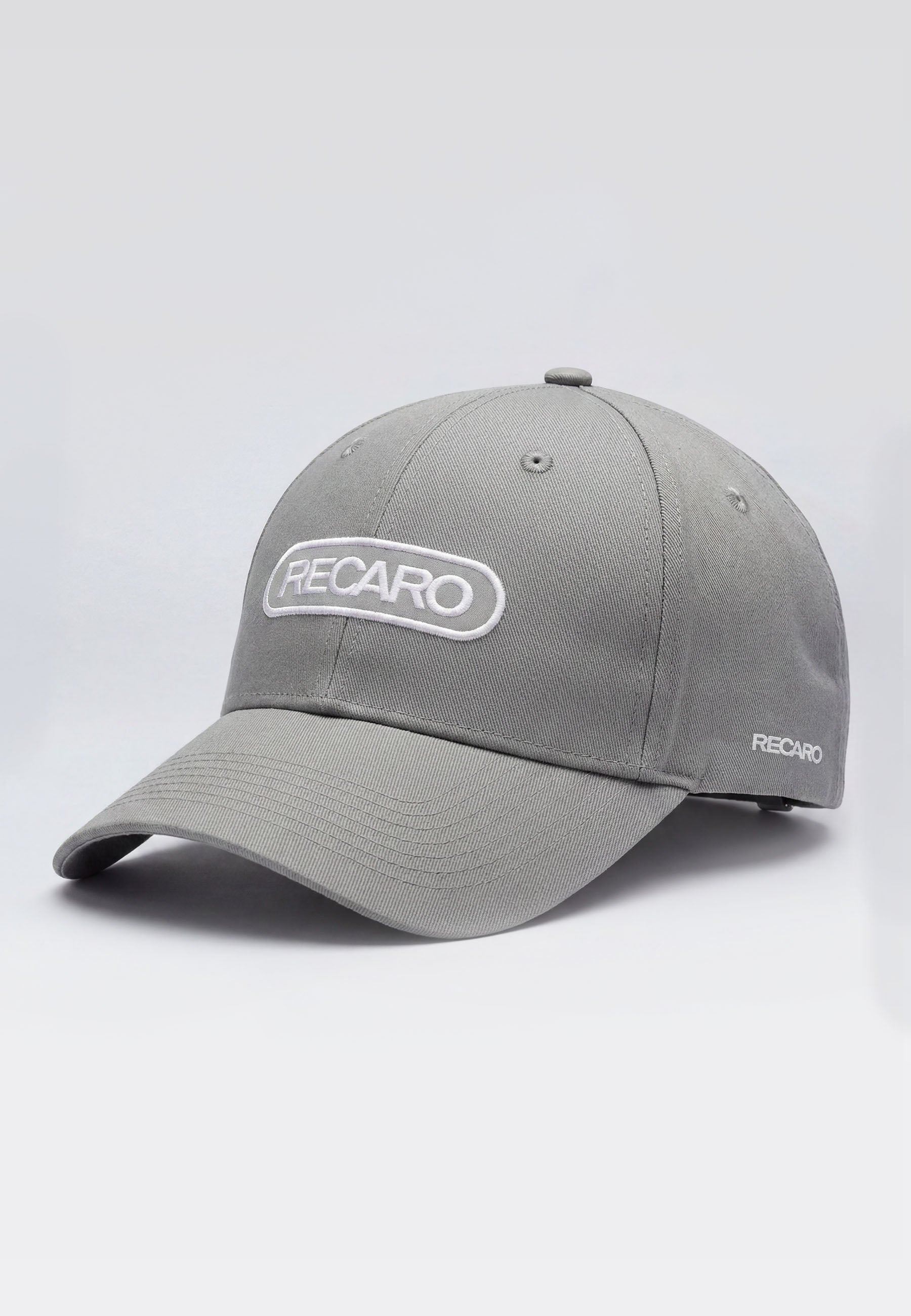 Die RECARO Shop Baseballcap Basic cool grey hat eine gebogene Krempe, 100% Baumwollmaterial, einen verstellbaren Verschluss und prominente weiße RECARO Logos auf der Vorderseite und an der Seite, die auf hellgrauem Hintergrund abgebildet sind.