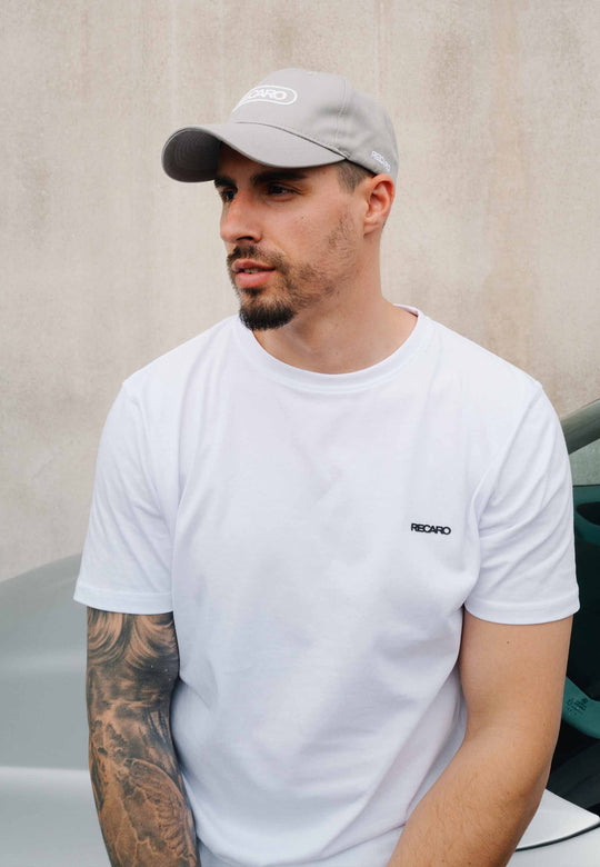Ein Mann mit kurzem Bart und Schnurrbart trägt eine RECARO Shop Baseball Cap Basic in Cool Grey und ein weißes T-Shirt mit einem kleinen RECARO-Logo. Er hat ein Tattoo am linken Arm und steht vor einer schlichten Wand und einem grauen Auto.