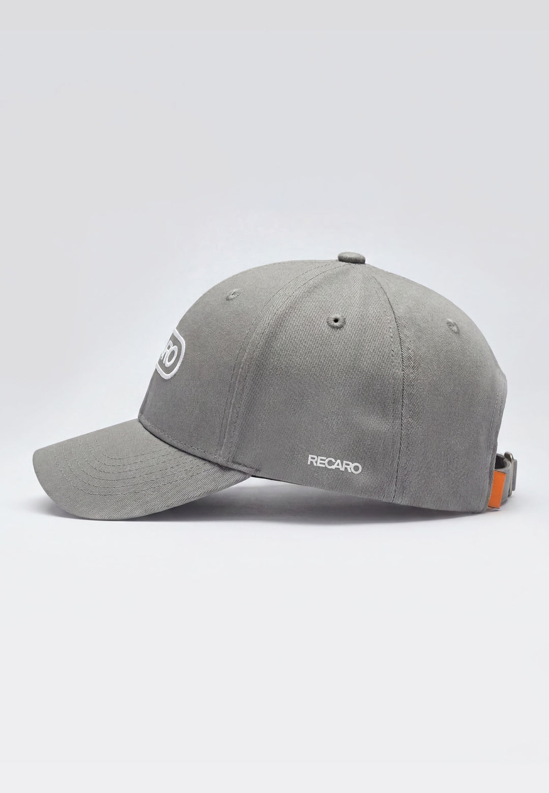 Die Baseball Cap Basic cool grey von RECARO Shop besteht aus 100% Baumwolle, hat einen gebogenen Schirm, ein weißes RECARO-Logo seitlich, einen verstellbaren Metallverschluss und ein kleines orangenes Detail nahe dem Verschluss.
