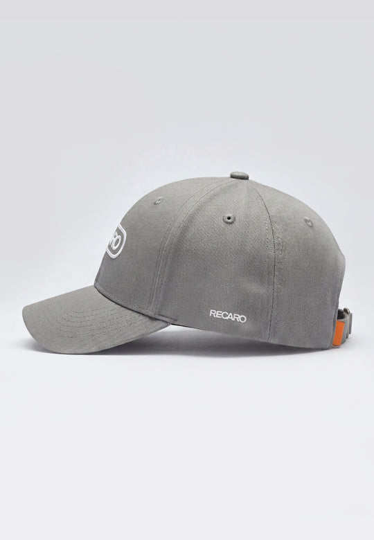 Die Baseball Cap Basic cool grey von RECARO Shop besteht aus 100% Baumwolle, hat einen gebogenen Schirm, ein weißes RECARO-Logo seitlich, einen verstellbaren Metallverschluss und ein kleines orangenes Detail nahe dem Verschluss.