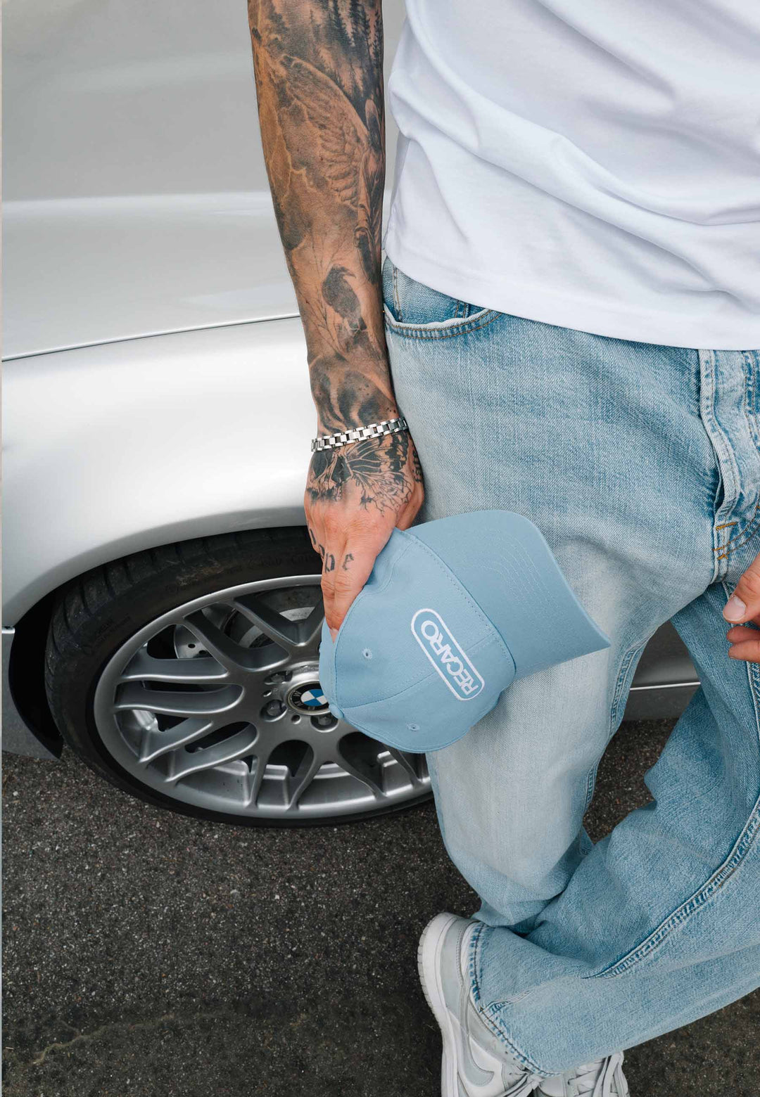 Eine Person in hellblauen Jeans, einem weißen Hemd und weißen Sneakers steht neben einem silbernen Auto und hält die RECARO Shop Baseball Cap Basic in der Farbe „Sky“ in der Hand. Ihr tätowierter Arm und ihre silbernen Armbänder sind zu sehen, im Hintergrund sind das Autorad und der Asphalt zu erkennen.