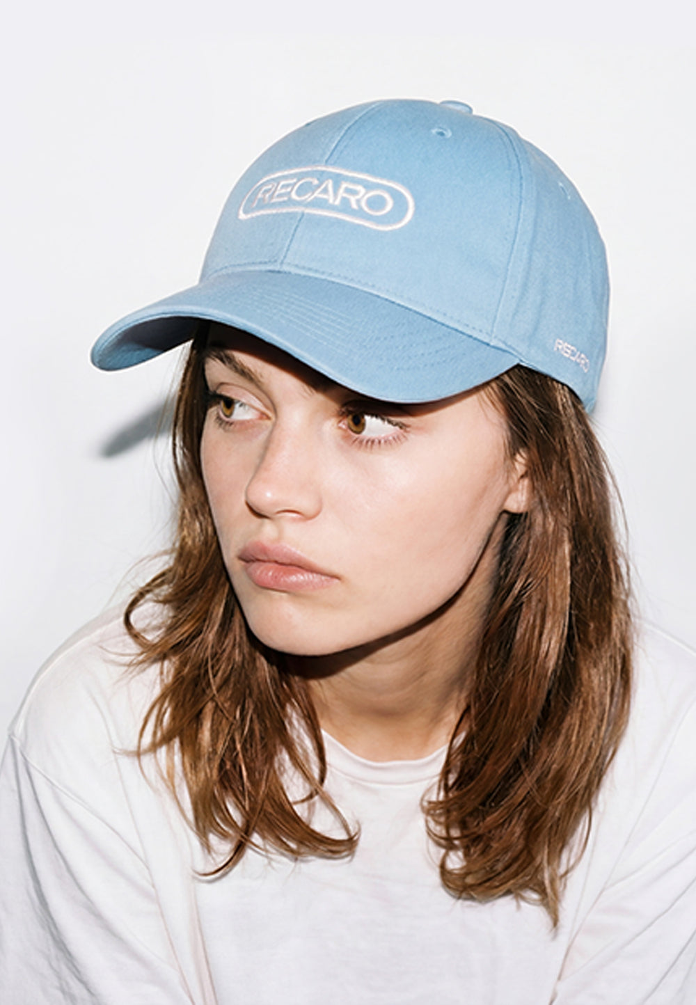 Eine junge Frau mit langen braunen Haaren trägt eine hellblaue RECARO Shop Baseball Cap Basic sky und ein weißes Hemd. Sie schaut leicht nach links und hat einen neutralen Gesichtsausdruck vor einem schlichten Hintergrund.