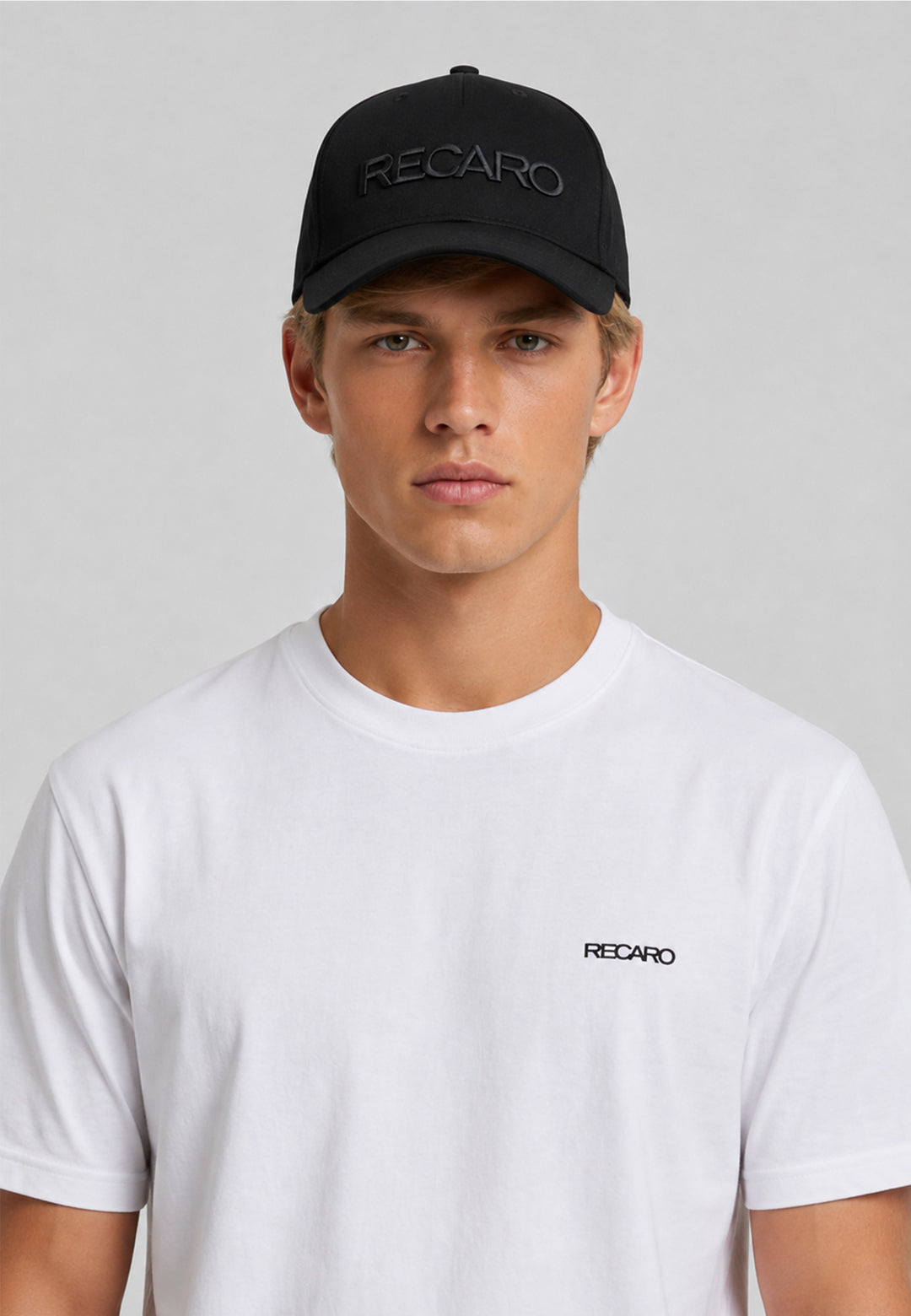Ein junger Mann, der eine schwarze „Recaro_Merch_Daniel Baseball Cap Originals“ mit 3D-Stickerei und Druckknopfverschluss sowie ein weißes RECARO-T-Shirt trägt, steht vor einem schlichten hellgrauen Hintergrund und blickt mit ausdruckslosem Gesicht direkt in die Kamera.
