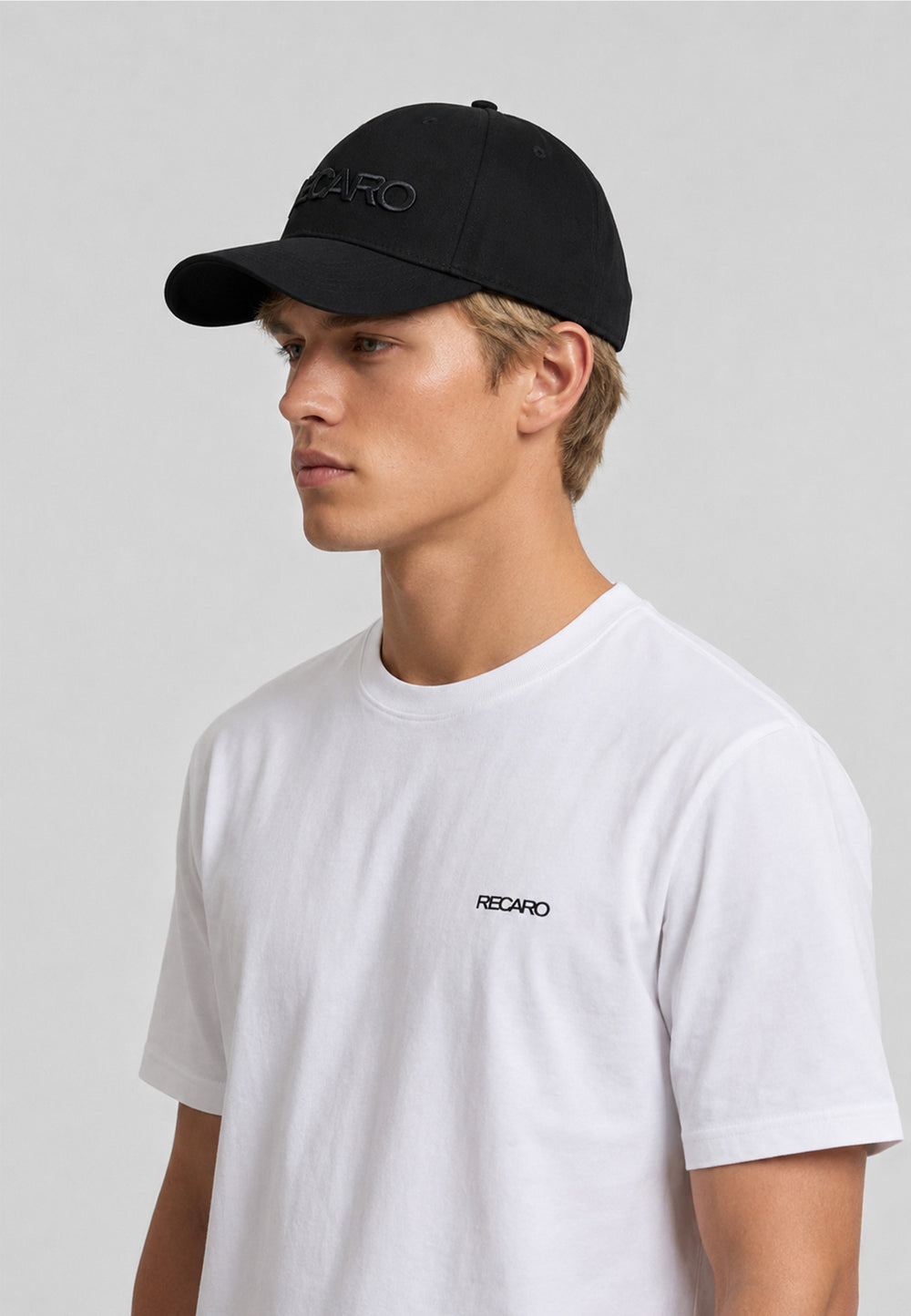 Ein junger Mann, der die Baseballkappe „Recaro_Merch_Daniel Baseball Cap Originals“ mit 3D-Stickerei sowie ein weißes T-Shirt mit dem Aufdruck „RECARO“ trägt, steht vor einem schlichten hellgrauen Hintergrund und blickt mit ausdruckslosem Gesicht zur Seite.