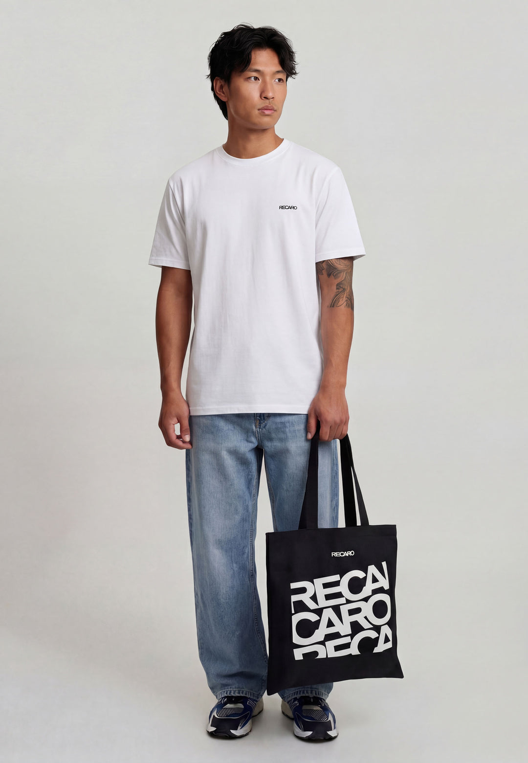 Ein Mann mit kurzen dunklen Haaren und einer Tätowierung auf dem linken Unterarm hält die RECARO Shop Baumwolltasche RECARO in der Hand, eine schwarze Baumwolltasche mit dem fetten weißen Schriftzug "RECARO". Er trägt ein schlichtes weißes T-Shirt, eine dunkle Hose und Turnschuhe.