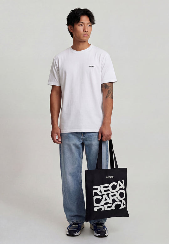 Ein Mann mit kurzen dunklen Haaren und einer Tätowierung auf dem linken Unterarm hält die RECARO Shop Baumwolltasche RECARO in der Hand, eine schwarze Baumwolltasche mit dem fetten weißen Schriftzug "RECARO". Er trägt ein schlichtes weißes T-Shirt, eine dunkle Hose und Turnschuhe.