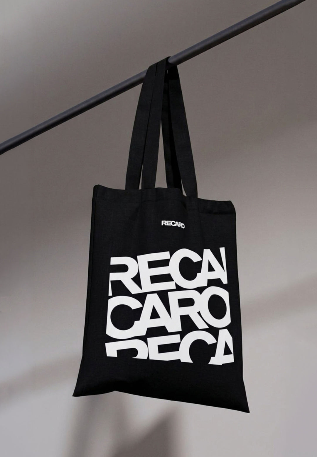 Die Baumwolltasche RECARO aus dem RECARO Shop hat einen auffälligen weißen Stapelschriftzug und hängt an einer schwarzen Stange vor einem grauen Hintergrund.