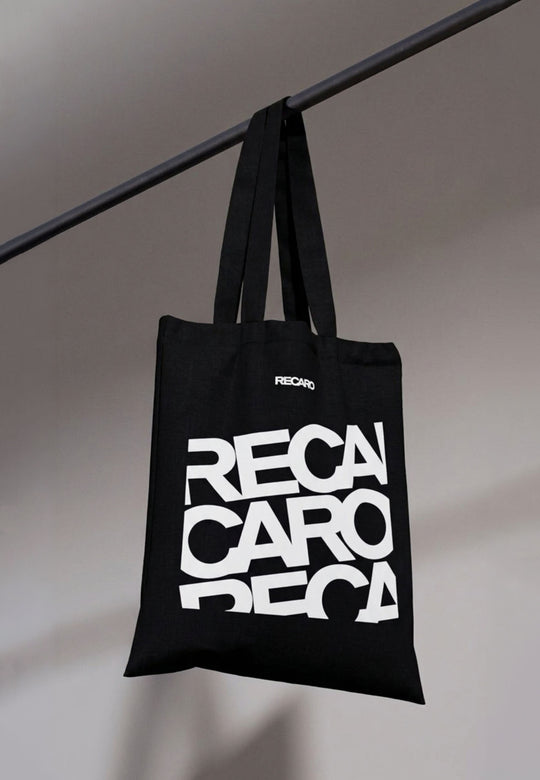 Die Baumwolltasche RECARO aus dem RECARO Shop hat einen auffälligen weißen Stapelschriftzug und hängt an einer schwarzen Stange vor einem grauen Hintergrund.