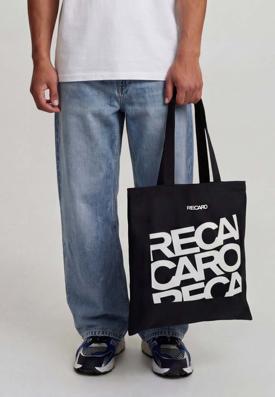 Eine Person hält die Baumwolltasche RECARO aus dem RECARO Shop mit dem fetten weißen Schriftzug "RECARO" in der Hand. Zu sehen ist nur die untere Hälfte der Person, die mit einer schwarzen Hose, einem weißen Hemd und blauen Sneakers bekleidet ist.