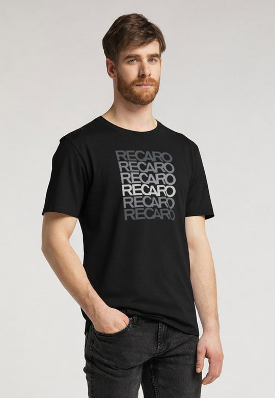Ein Mann mit kurzen braunen Haaren und einem Bart trägt das schwarze Recaro_Merch_Daniel T-Shirt Spektrum im Retrostil, mit wiederholten grauen Markenaufdrucken. Er steht mit einer Hand in der Tasche vor einem schlichten hellen Hintergrund. Hergestellt aus weicher Baumwolle.