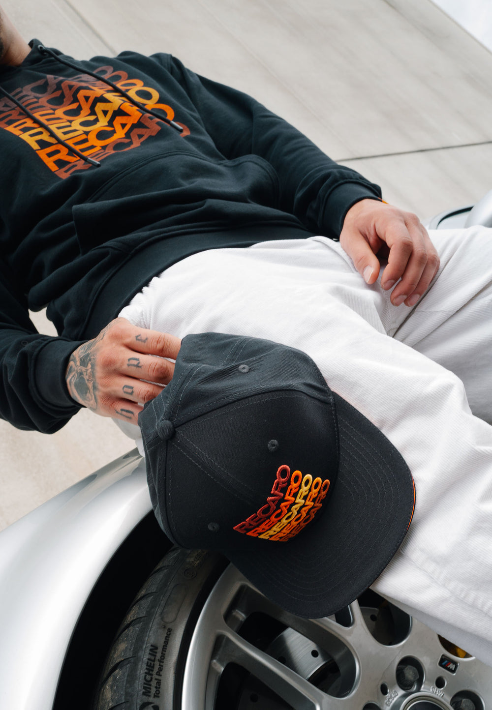 Eine Person im RECARO Shop Spektrum-Bundle – schwarzer Hoodie und dazu passende Snapback-Kappe – mit orange-gelben Grafiken trägt eine weiße Hose und lehnt an einem silbernen Auto, wobei Tattoos auf ihren Fingern zu sehen sind.