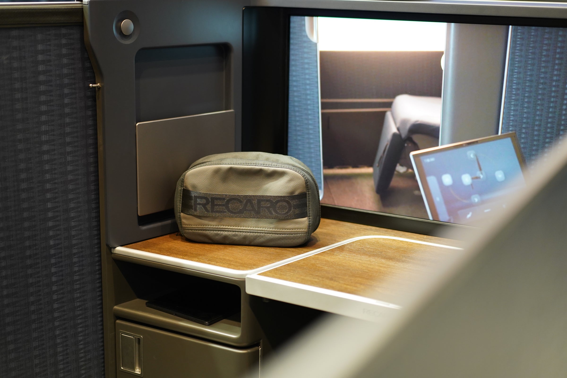 Eine kleine, graue Tasche mit dem RECARO-Logo steht auf einem Holztablett in einer modernen Flugzeug- oder Zugsuite, im Hintergrund befinden sich eine Trennwand, ein Stauraum und ein Fenster oder ein Bildschirm.
