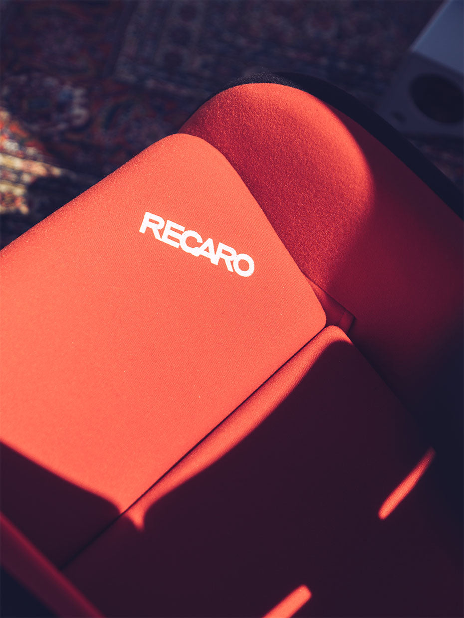 Nahaufnahme eines roten RECARO-Autositzes. Sonnenlicht wirft Schatten auf den Stoff und zeigt den Markennamen in weißen Buchstaben auf dem oberen Teil des Sitzes.