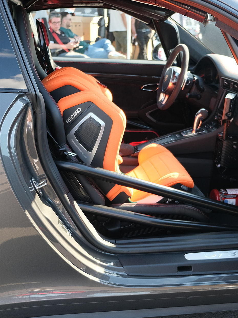 Nahaufnahme des Fahrzeuginnenraums mit leuchtend orangefarbenem Recaro-Rennsitz, Überrollkäfig und Feuerlöscher. Die Fahrertür ist geöffnet, im Hintergrund sind Personen außerhalb des Fahrzeugs zu sehen.