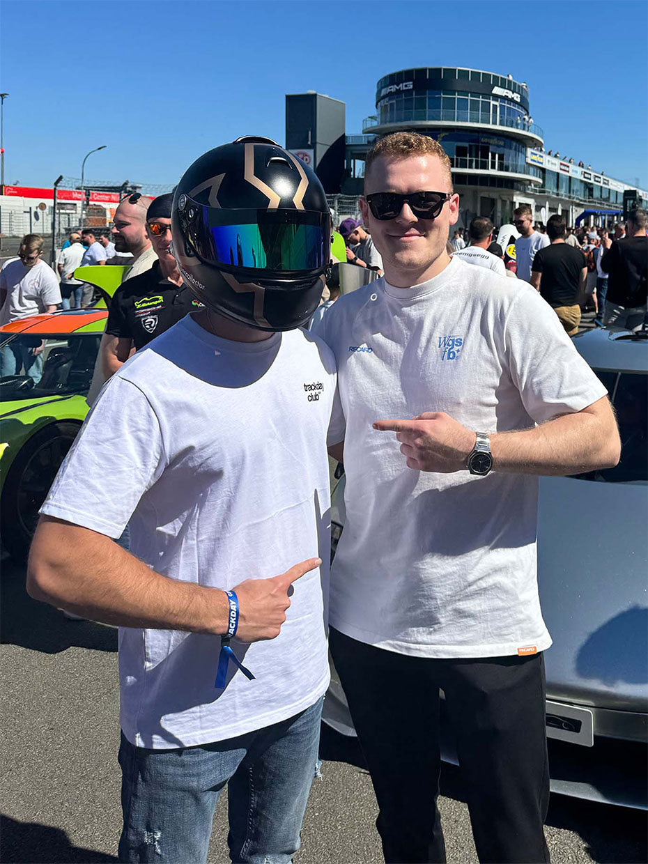 Zwei Männer posieren gemeinsam bei einer Autoveranstaltung. Einer trägt einen schwarzen Helm mit reflektierendem Visier und ein weißes T-Shirt, der andere eine Sonnenbrille und ein weißes Hemd. Beide lächeln und zeigen aufeinander. Hinter ihnen sind Menschenmengen und Sportwagen zu sehen.