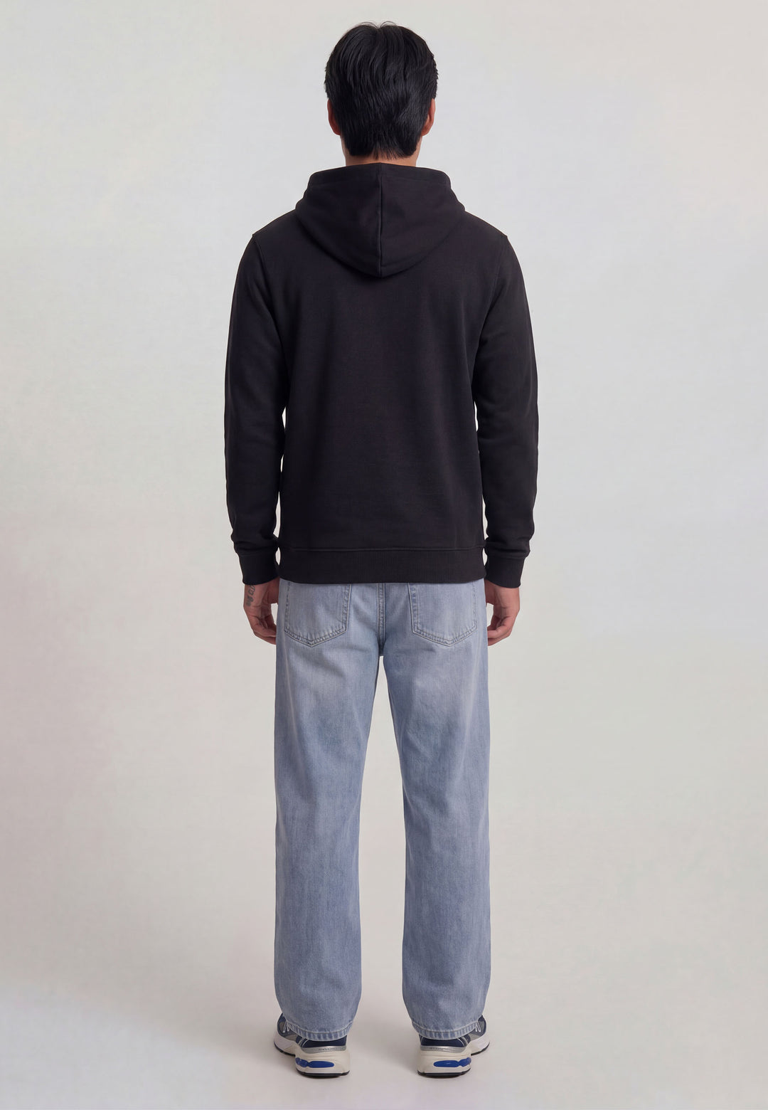 Eine Person mit kurzen dunklen Haaren blickt von der Kamera weg und trägt einen lässigen RECARO Shop Hoodie Originals in Schwarz, hellblaue Jeans und weiße Turnschuhe mit blauen Akzenten vor einem schlichten hellen Hintergrund.