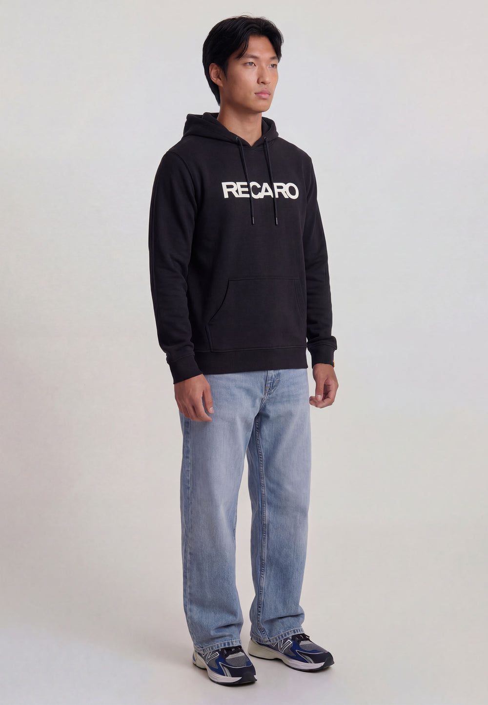 Jemand trägt einen lässig geschnittenen, unisex RECARO Shop Hoodie Originals, gestylt mit hellblauen Jeans und marineblauen Turnschuhen, vor einem schlichten hellen Hintergrund.
