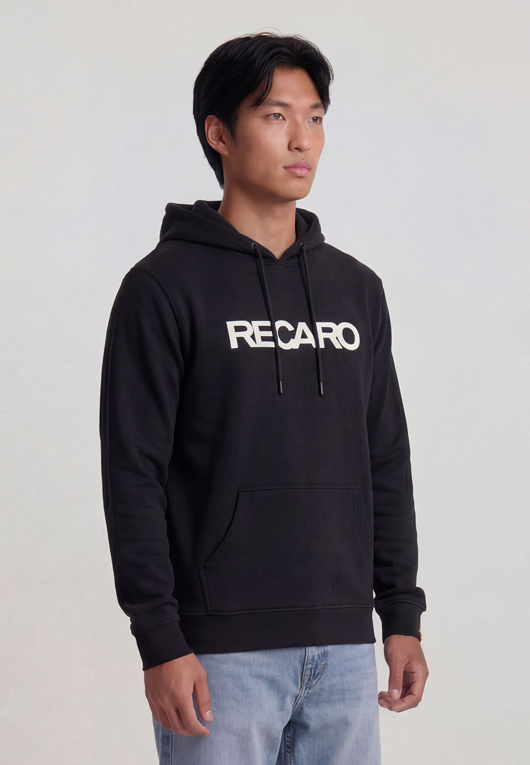 Ein Mann steht mit dem Gesicht nach vorne, die Hände an den Seiten, und trägt den lässigen RECARO Shop Hoodie Originals in unisex schwarz, gepaart mit einer hellblauen Jeans vor einem schlichten, hellen Hintergrund.