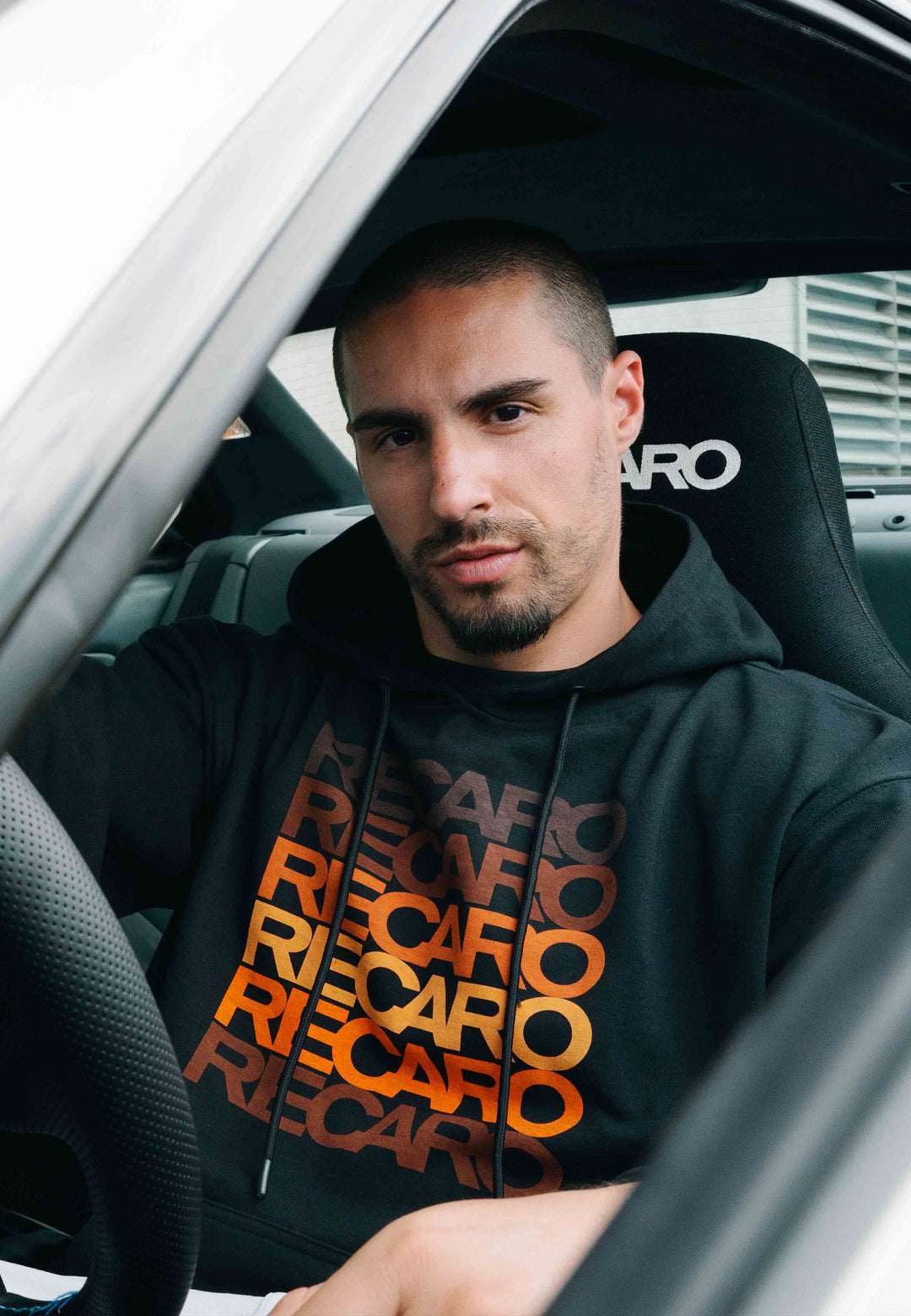 Ein Mann mit kurzen Haaren und einem gepflegten Bart sitzt auf dem Fahrersitz eines Autos und trägt den „Recaro_Merch_Daniel Hoodie Spektrum“ in Schwarz, einen Unisex-Hoodie im Retro-Look, auf dessen Vorderseite ein farbenfrohes Logo mehrfach aufgedruckt ist.